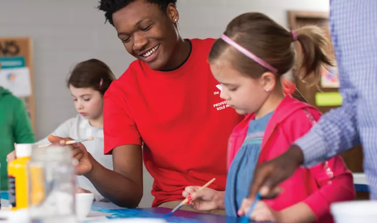 Young girl paints a picture while her teacher watches on.