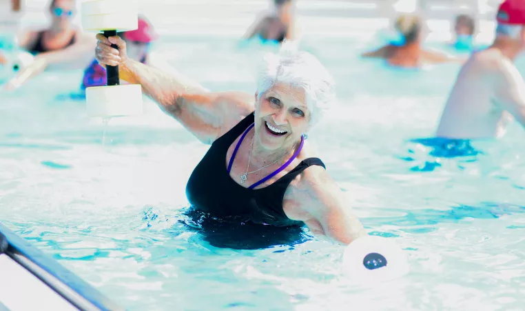 Older woman smiles and poses in the pool while holding water weights.