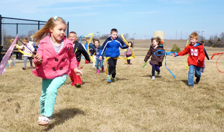 A group of young kids run and wave ribbons outside during their child care program.