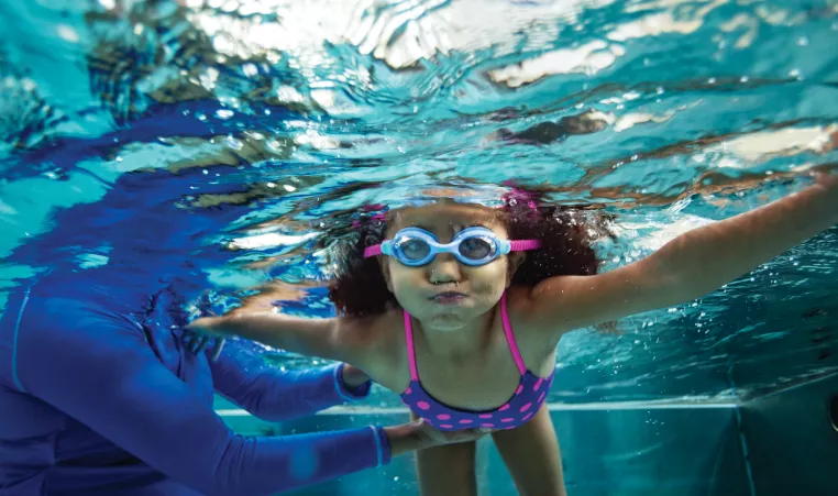 Girl holds her breath while her teacher helps her practice swimming under water.