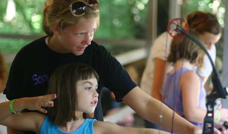 Photo of a camp counselor teaching a young girl how to hold an archery bow