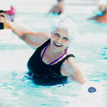 Older woman smiles and poses in the pool while holding water weights.